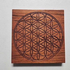 May include: A wooden coaster with a laser-etched geometric pattern, known as the Flower of Life, in a dark brown color.