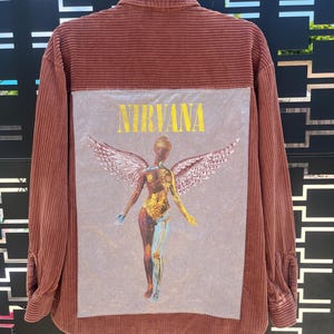 Może przedstawiać: Koszulka sztruksowa w kolorze rdzy z dużą grafiką na plecach. Grafika przedstawia słowo "NIRVANA" w kolorze żółtym nad stylizowaną postacią ze skrzydłami. Postać to mieszanka złota, niebieskiego i odcieni skóry.