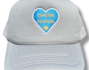 May include: A gray trucker hat with a blue heart-shaped patch that says "Coastal Cowgirl" in gold lettering. The patch also features a small gold seashell.