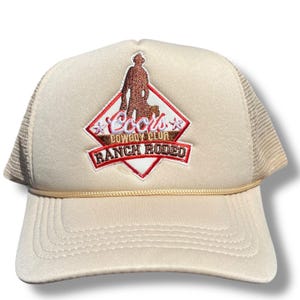 May include: A beige trucker hat with a brown and red embroidered patch that says "Cool's Cowboy Club Ranch Rodeo" with a silhouette of a cowboy and a dog.