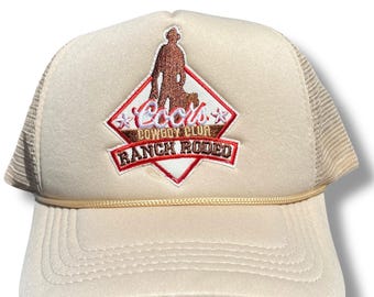 May include: A beige trucker hat with a brown and red embroidered patch that says "Cool's Cowboy Club Ranch Rodeo" with a silhouette of a cowboy and a dog.