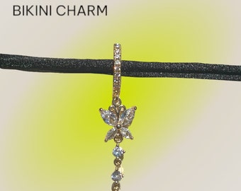 May include: A gold-colored removable bikini charm with a butterfly design. The butterfly is made of clear, sparkling stones. Below the butterfly are four smaller, round, clear stones. The text "REMOVABLE BIKINI CHARM" is at the top.