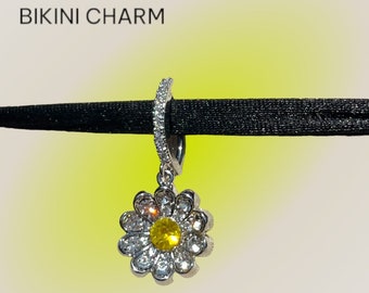 May include: A silver charm in the shape of a daisy with a yellow center and clear rhinestones. The charm is attached to a silver ring, which is designed to be attached to a bikini.