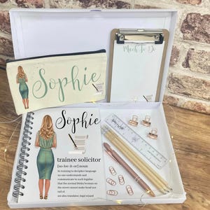 May include: A white gift box with a white notebook, a pencil case, a clipboard, and a ruler. The notebook and pencil case have a design of a woman in a green dress and the name "Sophie" on them. The clipboard has the words "Much to Do" written on it. The ruler is clear plastic with the words "Jane & Wright" printed on it. The box also contains a set of gold clips, pencils, and a pen.