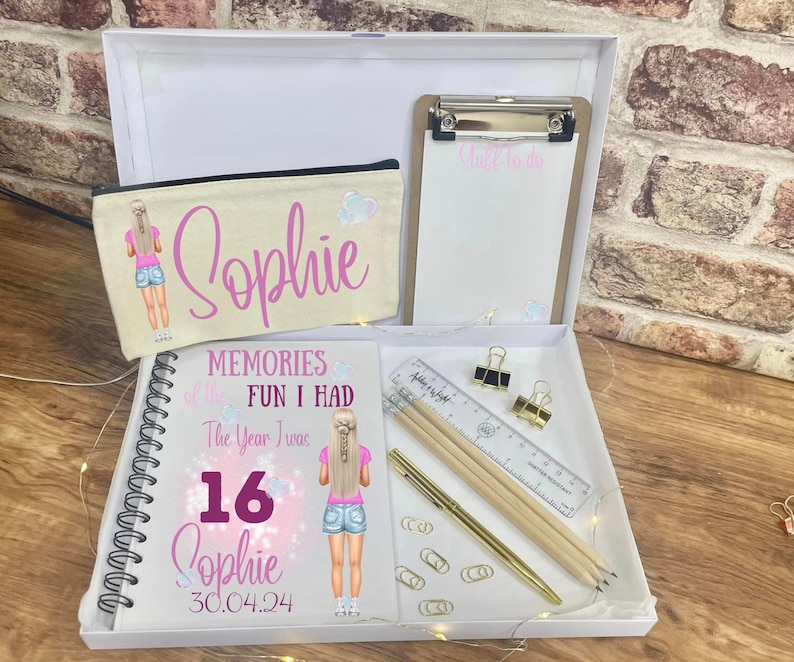 May include: A white gift box containing a personalized pencil case, a spiral-bound notebook, a clipboard, and stationery. The pencil case and notebook feature the name "Sophie" and a cartoon illustration. The notebook reads "Memories of the Fun I Had The Year I was 16 Sophie 30.04.24".