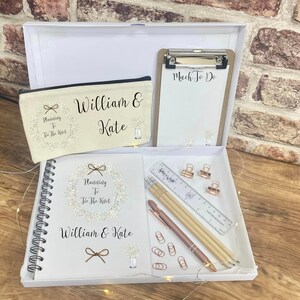 May include: A white gift box containing a cream-colored canvas pencil case, a clipboard with "Much To Do" text, a spiral-bound notebook with "Planning To Tie The Knot" and "William & Kate" text, a clear ruler, two wooden pencils, a pen, and rose gold clips.