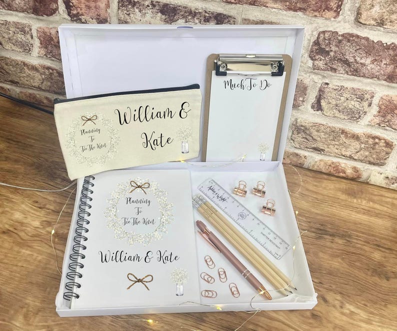 May include: A white gift box containing a cream-colored canvas pencil case, a clipboard with "Much To Do" text, a spiral-bound notebook with "Planning To Tie The Knot" and "William & Kate" text, a clear ruler, two wooden pencils, a pen, and rose gold clips.