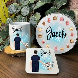 May include: A white ceramic mug, coaster, and round tin featuring the name "Jacob" in cursive. The mug and coaster have a graphic of a person in a blue uniform holding a coffee cup. The tin is decorated with cookie characters.