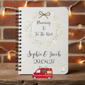 May include: A white spiral-bound notebook featuring a printed wreath of white baby's breath flowers with a brown twine bow at the top. The text reads Planning To Tie The Knot, Sophie & Jacob, and the date 29.04.27, with a small floral illustration.