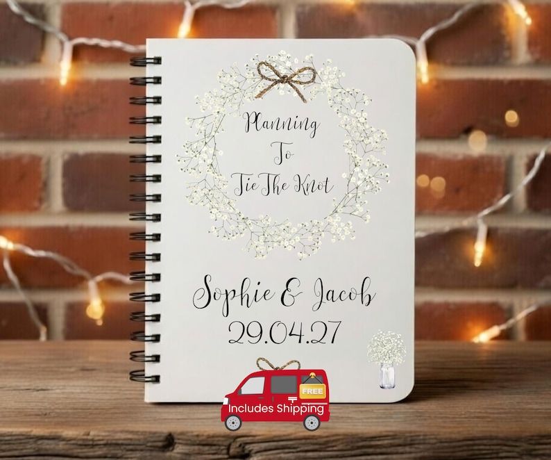 May include: A white spiral-bound notebook featuring a printed wreath of white baby's breath flowers with a brown twine bow at the top. The text reads Planning To Tie The Knot, Sophie & Jacob, and the date 29.04.27, with a small floral illustration.