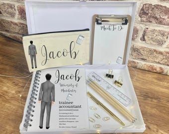 May include: A white gift box with a white background. The box contains a white notebook with a black spiral binding, a pencil case with a black and white illustration of a person, a clipboard with a white notepad, a pen, pencils, a ruler, and paper clips. The notebook has the name "Jacob" and the text "University of Manchester" printed on it. The pencil case has the name "Jacob" printed on it. The clipboard has the text "Much To Do" printed on it.