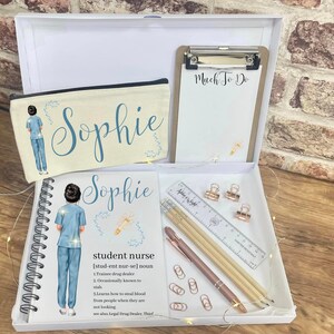 May include: A white gift box with a blue and white pencil case, a blue and white spiral notebook, a clipboard with the text "Much To Do", and a ruler, pencils, and pens. The pencil case and notebook have the name "Sophie" printed on them. The notebook also has the text "student nurse" and a definition of the term.