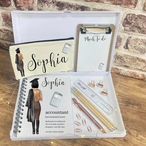 May include: A white gift box containing a pencil case, notepad, clipboard, and stationery. The pencil case and notebook feature the name "Sophia" and an illustration of a graduate. The clipboard reads "Much To Do". Includes pencils, a ruler, paper clips, and a pen.
