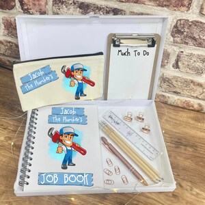 May include: A white gift box containing a pencil case, clipboard, notebook, pens, ruler, and paper clips. The pencil case and notebook feature a cartoon plumber graphic and the text "Jacob The Plumber's". The clipboard has the text "Much To Do".