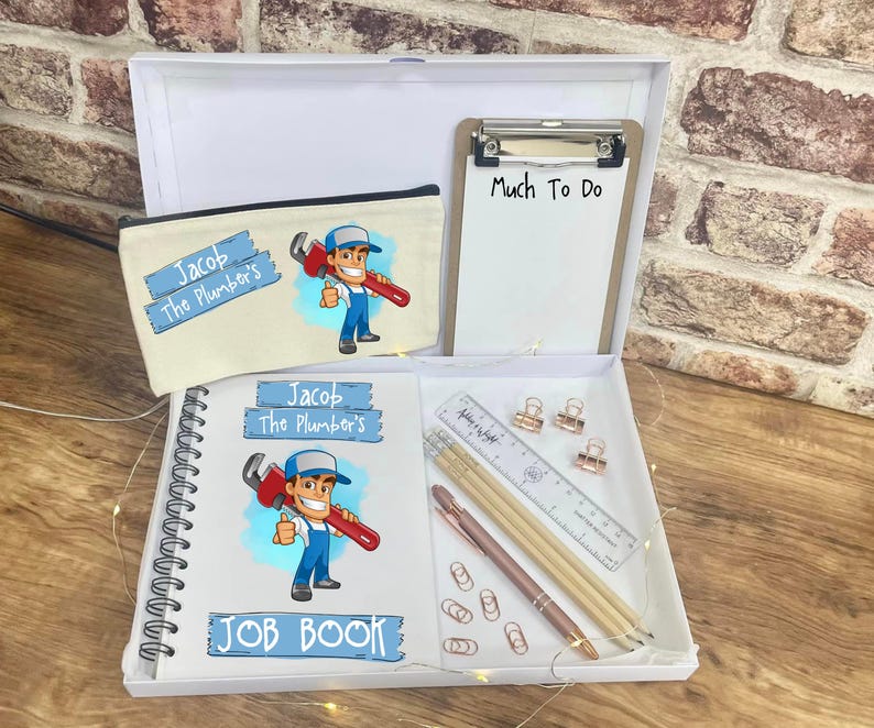 May include: A white gift box containing a pencil case, clipboard, notebook, pens, ruler, and paper clips. The pencil case and notebook feature a cartoon plumber graphic and the text "Jacob The Plumber's". The clipboard has the text "Much To Do".