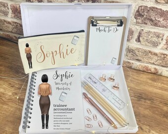 May include: A white gift box with a white background. The box contains a white notebook with a gold spiral binding, a white pencil case with a gold zipper, a white clipboard with a gold clip, and a set of gold pencils, a ruler, and paper clips. The notebook and pencil case have the name "Sophie" printed on them in gold lettering. The clipboard has the words "Much To Do" written on it in black lettering. The notebook also has the words "University of Manchester" and "trainee accountant" printed on it in gold lettering.