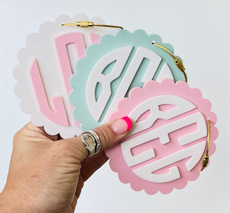 Puede incluir: Tres decoraciones de pared redondas con bordes festoneados, fondo blanco y monogramas rosa, azul claro y rosa oscuro. Los monogrammes son de estilo tradicional.