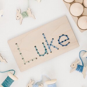 May include: A wooden board with the name "Luke" spelled out in blue and green yarn. The board is surrounded by wooden spools of yarn and wooden bunny shapes.