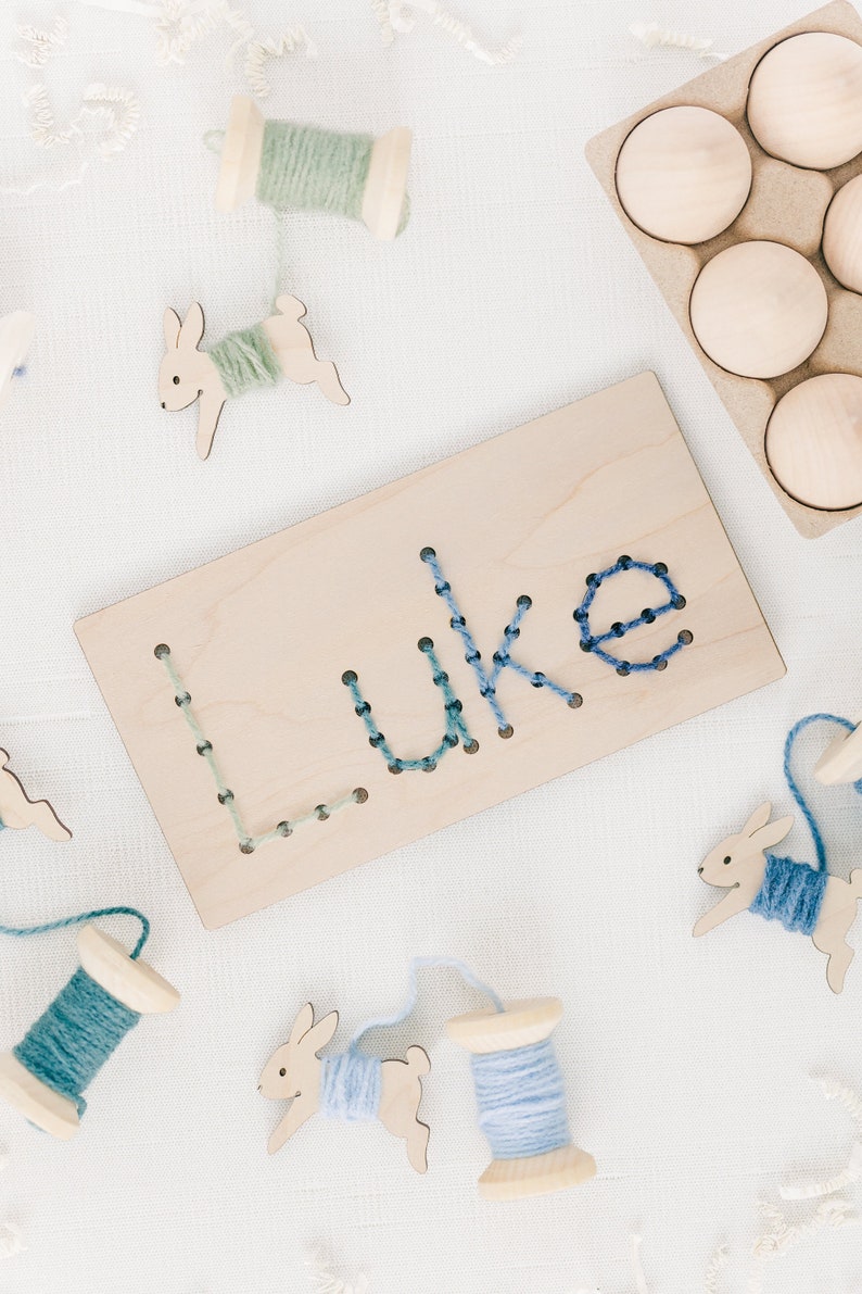 May include: A wooden board with the name "Luke" spelled out in blue and green yarn. The board is surrounded by wooden spools of yarn and wooden bunny shapes.