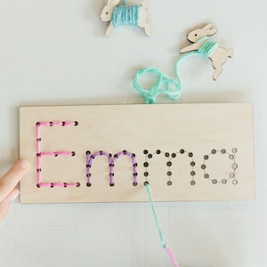 May include: A wooden board with the name "Emma" spelled out in holes. Pink, purple, and teal yarn is threaded through the holes. Two wooden bunny shapes with yarn attached are in the background.