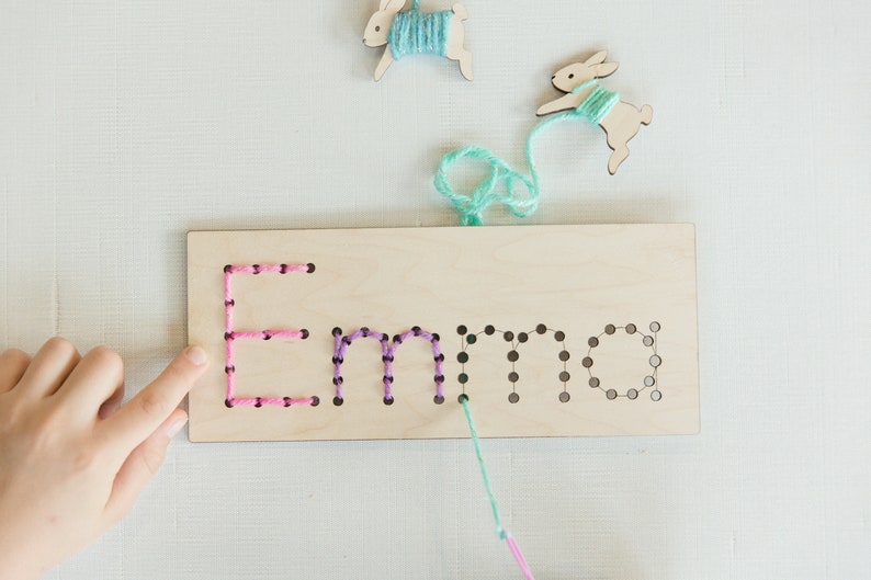 May include: A wooden board with the name "Emma" spelled out in holes. Pink, purple, and teal yarn is threaded through the holes. Two wooden bunny shapes with yarn attached are in the background.