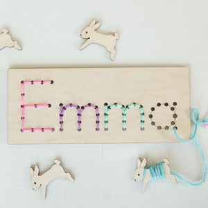 May include: Wooden board with the name "Emma" spelled out in holes. The holes are filled with colorful yarn. There are four wooden bunny shapes around the board, one with yarn attached.