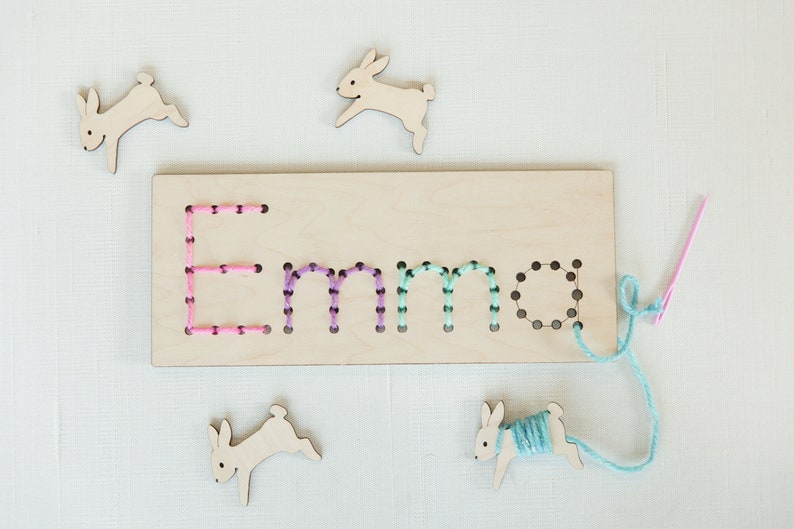 May include: Wooden board with the name "Emma" spelled out in holes. The holes are filled with colorful yarn. There are four wooden bunny shapes around the board, one with yarn attached.