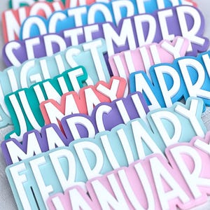 May include: A collection of colorful month name labels in various pastel shades, including pink, blue, and green. Each label features a white, bold font. The labels are arranged in a layered, slightly angled pattern.
