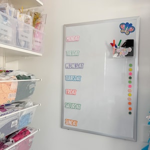 Puede incluir: Una pizarra blanca de borrado en seco con un marco plateado. La pizarra tiene una pegatina de mariposa colorida, un recipiente transparente con marcadores y una columna de pegatinas coloridas con los d&iacute;as de la semana escritos en ellas. Los d&iacute;as de la semana son: lunes, martes, mi&eacute;rcoles, jueves, viernes, s&aacute;bado y domingo.
