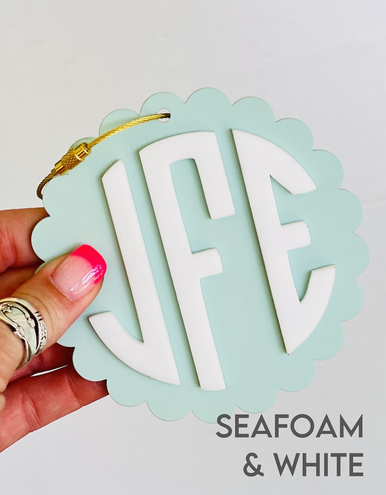 Puede incluir: Una etiqueta de equipaje azul claro con un monograma blanco "JFE" y una cadena dorada. La etiqueta tiene un borde festoneado y el texto "SEAFOM & WHITE" est&aacute; impreso debajo del monograma.
