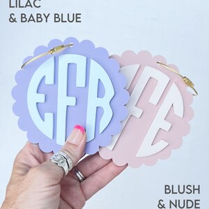 Puede incluir: Dos etiquetas de equipaje redondas, festoneadas y personalizadas con anillos de metal dorado. La etiqueta de la izquierda es lila con un monograma azul beb&eacute; "EFR". La etiqueta de la derecha es ruborizada con un monograma nude "ME".