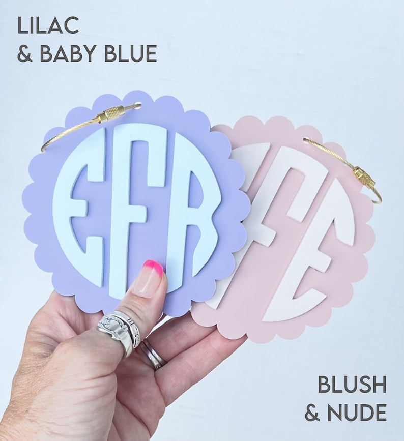 Puede incluir: Dos etiquetas de equipaje redondas, festoneadas y personalizadas con anillos de metal dorado. La etiqueta de la izquierda es lila con un monograma azul beb&eacute; "EFR". La etiqueta de la derecha es ruborizada con un monograma nude "ME".