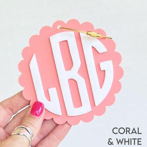Puede incluir: Etiqueta de equipaje de color coral y blanco con el monograma "L.B.G." en letras blancas. La etiqueta tiene un borde festoneado y un lazo de metal dorado.