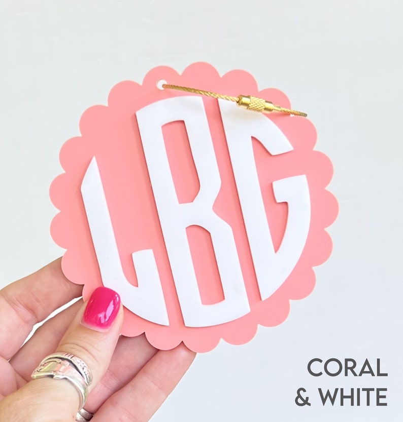 Puede incluir: Etiqueta de equipaje de color coral y blanco con el monograma "L.B.G." en letras blancas. La etiqueta tiene un borde festoneado y un lazo de metal dorado.
