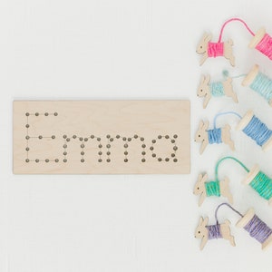 May include: Wooden board with the name "Emma" spelled out in holes.  Five wooden bunny figures with yarn wrapped around them, each a different color: pink, blue, light blue, green, and purple.