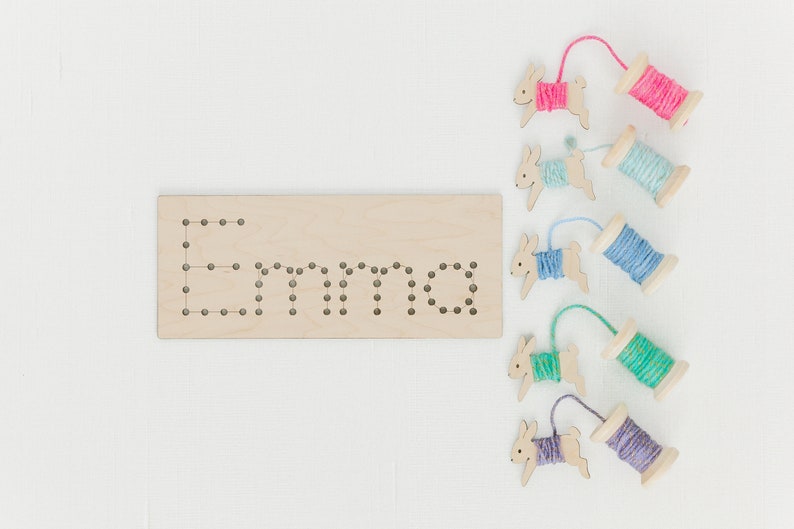 May include: Wooden board with the name "Emma" spelled out in holes.  Five wooden bunny figures with yarn wrapped around them, each a different color: pink, blue, light blue, green, and purple.