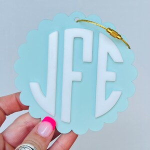Puede incluir: Una etiqueta de equipaje azul claro con un borde blanco festoneado y un monograma blanco "JFE" en un c&iacute;rculo. La etiqueta tiene un lazo de metal dorado y un cierre de metal dorado.