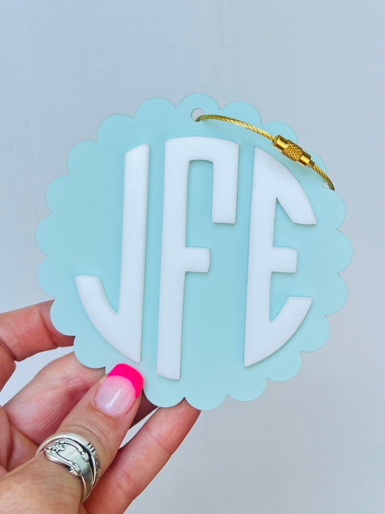 Puede incluir: Una etiqueta de equipaje azul claro con un borde blanco festoneado y un monograma blanco "JFE" en un c&iacute;rculo. La etiqueta tiene un lazo de metal dorado y un cierre de metal dorado.