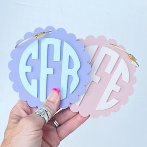 Peut inclure: Deux &eacute;tiquettes &agrave; bagages rondes et festonn&eacute;es avec des anneaux en m&eacute;tal dor&eacute;. L'&eacute;tiquette de gauche est lavande avec un monogramme bleu clair "EFR". L'&eacute;tiquette de droite est rose avec un monogramme blanc "ME".