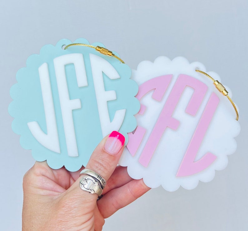 Puede incluir: Dos etiquetas de equipaje de acr&iacute;lico blanco, festoneadas, con anillos de metal dorado. Una etiqueta es azul claro con letras blancas que deletrean "JFE" y la otra etiqueta es blanca con letras rosas que deletrean "EFL".