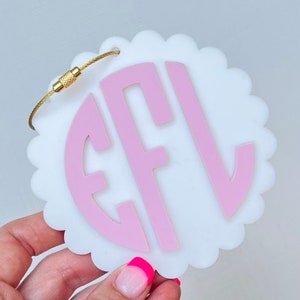 Puede incluir: Etiqueta de equipaje blanca con un monograma rosa "EFL" en un c&iacute;rculo. La etiqueta tiene un borde festoneado y un lazo de metal dorado.