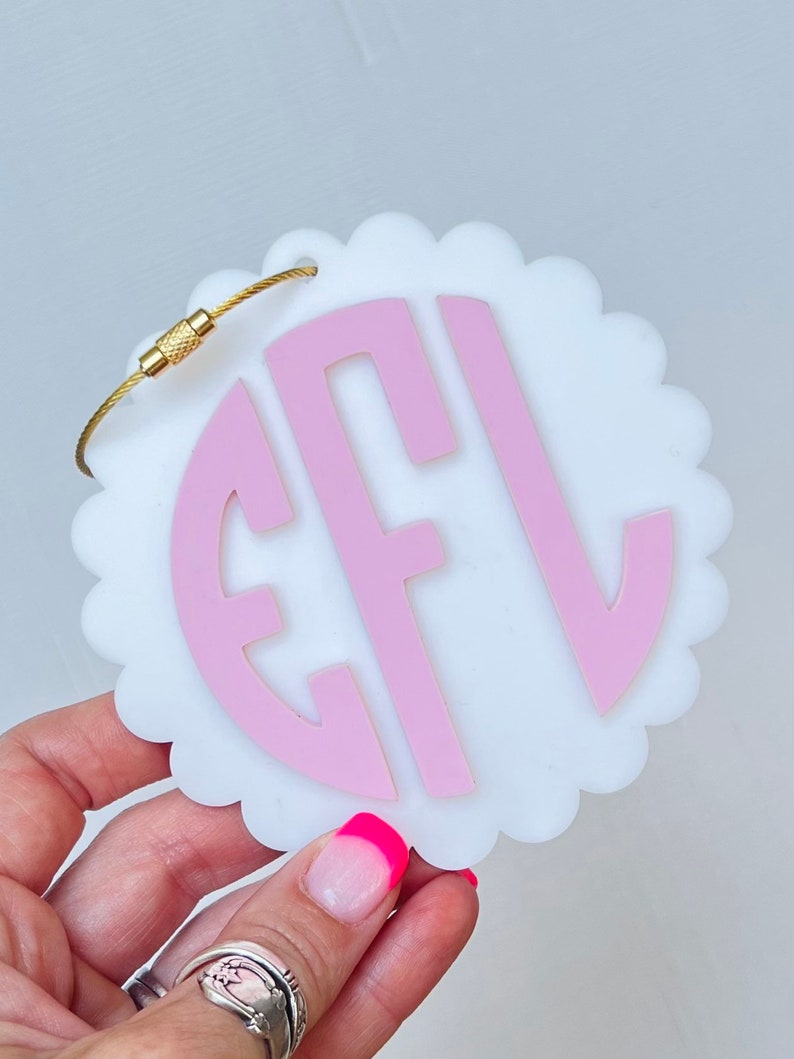 Puede incluir: Etiqueta de equipaje blanca con un monograma rosa "EFL" en un c&iacute;rculo. La etiqueta tiene un borde festoneado y un lazo de metal dorado.