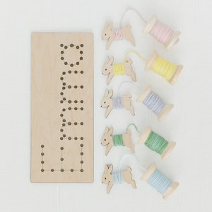May include: A wooden board with the word "LULA" spelled out in holes, and five wooden bunny shapes with yarn spools attached. The yarn spools are in pink, yellow, purple, green, and blue.