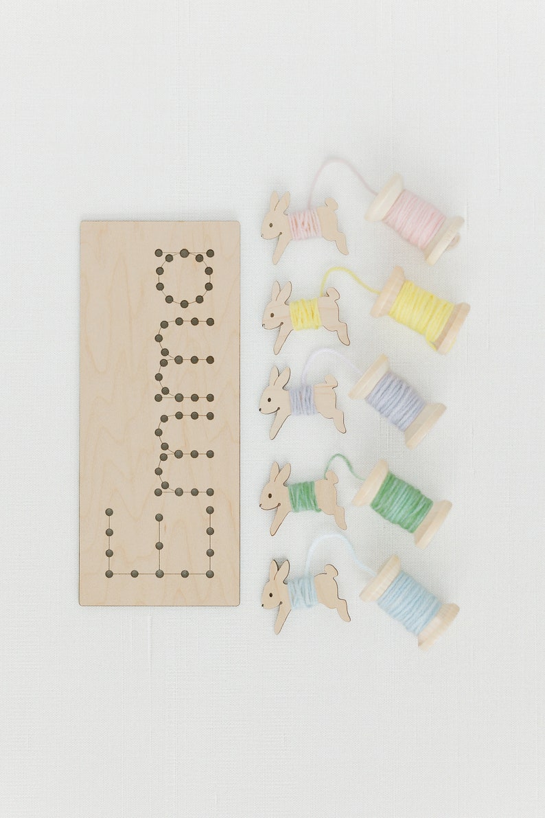 May include: A wooden board with the word "LULA" spelled out in holes, and five wooden bunny shapes with yarn spools attached. The yarn spools are in pink, yellow, purple, green, and blue.