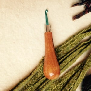 May include: A wooden crochet hook with a green metal hook. The hook is lying on a white surface with green yarn.