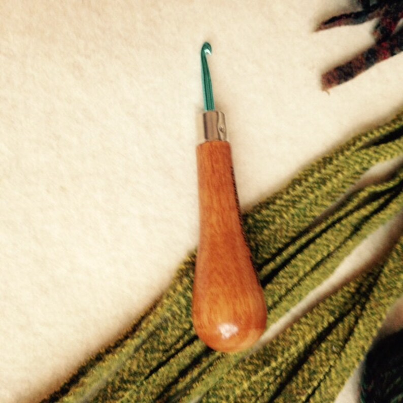 May include: A wooden crochet hook with a green metal hook. The hook is lying on a white surface with green yarn.