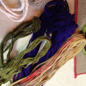 May include: A close-up of a variety of yarn and embroidery floss in different colors, including white, brown, green, purple, and beige. The yarn is arranged in a pile on a white surface.