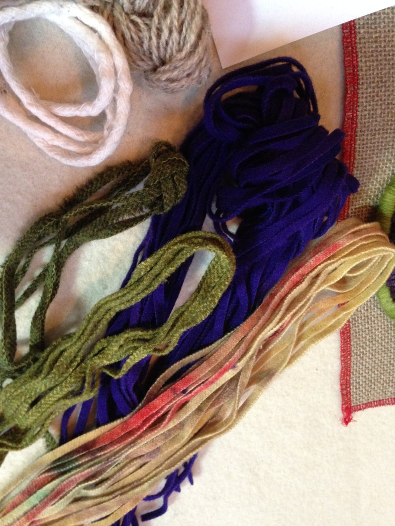 May include: A close-up of a variety of yarn and embroidery floss in different colors, including white, brown, green, purple, and beige. The yarn is arranged in a pile on a white surface.
