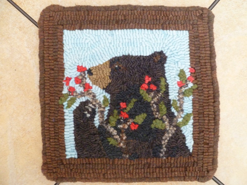 Pu&ograve; includere: Un tappeto di orso marrone e nero con una cornice marrone. L'orso guarda a sinistra e ha un cespuglio di bacche rosse dietro di lui.