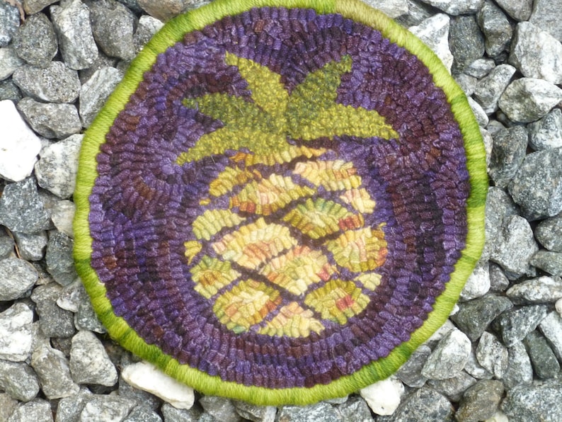 May include: A round, purple and green wool rug with a pineapple design. The pineapple is made of yellow, orange, and green wool.
